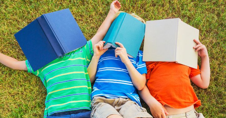 3 young boys laid on the grass with books over their faces
