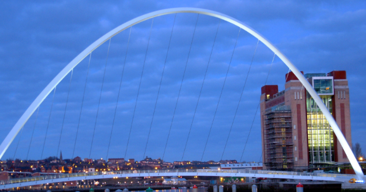 Close-up of Gateshead Millenium Bridge in the evening. Blue sky. Buildings with yellow lights behind.