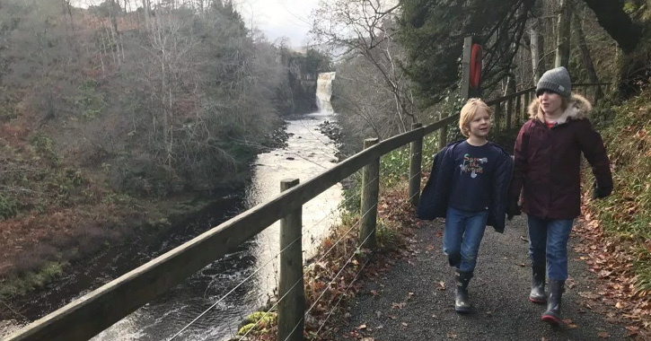 Two boys in winter clothing walking along the High Force Waterfall woodland trail with waterfall in background.