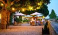 Outdoor area of the hotel at dusk. 
Several patio tables are dotted along the gravel with neutral parasols and lights hanging from the nearby trees.