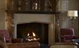 A roaring fire with burgundy comfortable chairs at either side. A tall lamp with a cream shade is behind the right hand chair.