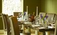 A long rectangular dining table set for dinner with wine glasses, white napkins, lit candles, purple flowers and a wine bucket.